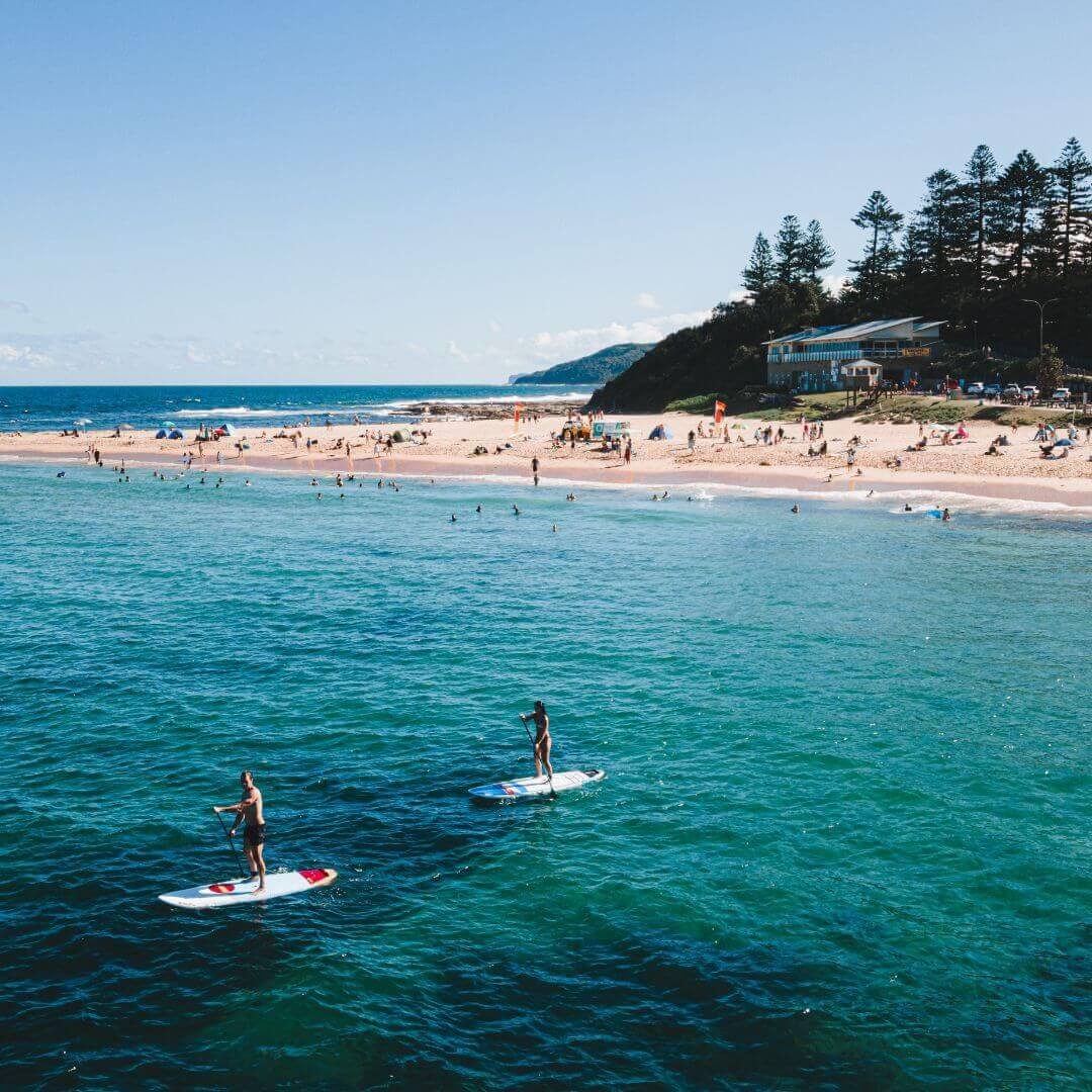 A calm blue sea with a beach in the background as two people paddle board along the water